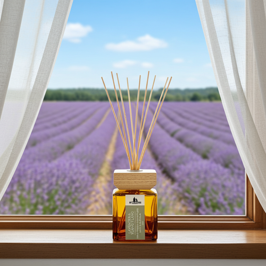A reed diffuser bottle with amber liquid and natural reed sticks sits on a wooden windowsill. Outside the window is a blurred lavender field under a blue sky, with white sheer curtains framing the scene.