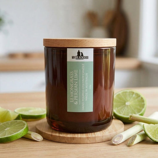 A glass jar candle with a light wood lid sits on a wooden coaster on a tabletop, surrounded by lime slices and lemongrass stalks. The candle has a green and white label that reads 'MY BLACK DOG' and 'LEMONGRASS & PERSIAN LIME'.